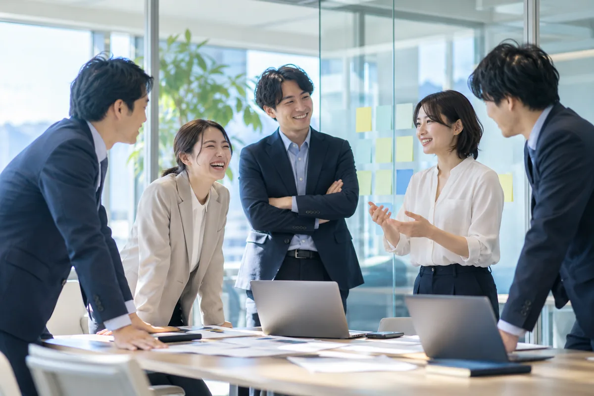 小さなプロジェクトルームで、若手のコンサルタント5人がテーブルを囲み、笑いながら話し合っている様子
